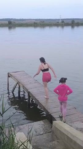 Two girls are on a wooden pier at a lake. The older girl in a black top and pink skirt jumps into the water, creating a large splash. A younger girl in a pink hoodie and pink leggings watches from the pier. Another person, partially obscured by reeds, is in the water holding a bag. The sky is overcast, and the water is calm. The pier extends over the water, with reeds growing along the bank.