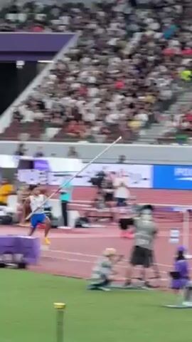 The video shows a pole vault attempt at a track and field event in a stadium packed with spectators. A pole vaulter wearing a white and blue uniform sprints towards the bar, plants their pole, vaults into the air, and attempts to clear the bar. The vaulter does not clear the bar and falls onto the landing pad below. Several officials stand by the landing pad observing the jump. Banners with sponsor logos are visible in the background.