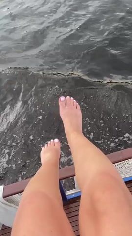 The video shows a person's legs and feet dangling over the side of what appears to be a boat. The water is dark and choppy. The person's toenails are painted a light pink color. The person's legs are bare and appear to be tanned. The video is filmed from a first-person perspective.