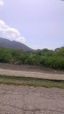 The video shows a moving shot from a vehicle. The view is of a road with a curb on the left side and some vegetation and hills in the background. The sky is partly cloudy. The time stamp indicates that this is the beginning of the video.