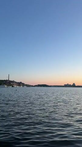 The video displays a wide, static shot of a body of water under a clear sky. The sky is light blue at the top, transitioning to a pale orange-pink at the horizon, indicative of twilight. The water is dark blue-grey with gentle ripples. On the distant left shore, a tall, slender chimney-like structure is prominent among other buildings and several boats. On the distant right shore, a cluster of larger, blocky buildings is visible. No people or specific actions are observed.