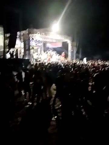 A night time outdoor concert is being performed on a stage. A large crowd of people are in front of the stage, facing the performers. The crowd is silhouetted against the stage lights and is actively engaged in the performance. The stage is set up with musical instruments and lighting rigs, and several performers are visible on stage. The atmosphere appears to be energetic and festive.