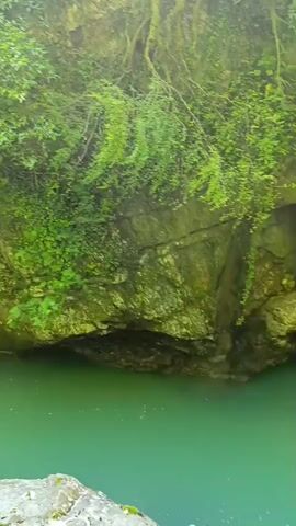 The video shows a river flowing through a canyon. The water is a bright turquoise color, and the canyon walls are covered in lush green vegetation. Sunlight streams down into the canyon, illuminating the water and the rocks. The camera pans across the scene, giving a sense of the depth and beauty of the natural landscape. There are no people visible in the video.