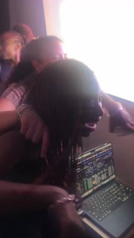 The video shows a group of people gathered around a laptop in what appears to be a DJ setup. At least three people are visible, their faces obscured by motion blur. One person is standing directly in front of the laptop, their hand hovering over the keyboard as if controlling the DJ software displayed on the screen. Another person, possibly two, are standing or leaning in from behind, their hands also near the laptop. The environment is dimly lit with a large, bright light source in the background, possibly a projector screen. The scene suggests a lively and interactive atmosphere, likely related to music or entertainment.