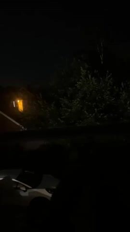 The video shows a dark outdoor scene at night. In the background, a house with a glowing orange window is visible. Dense green foliage is situated between the house and the foreground. In the foreground, the dark silhouette of a white car with its hood open is present. The lighting is dim, suggesting it is nighttime.