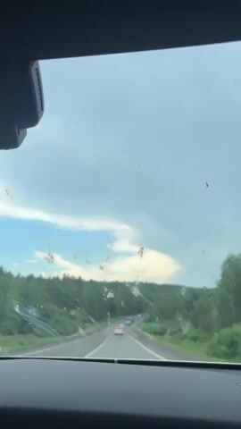 This is a video clip of a drive on a highway in Urten', Russia. The view is from inside a car, looking out through the windshield. The windshield is dirty, with numerous streaks and spots of dried mud or debris, which obscure the view slightly.

The scene depicts a two-lane highway curving to the right, surrounded by a dense forest of green trees on either side. The road ahead is visible with several cars driving in the same direction. The sky is overcast with dramatic, thick clouds, some of which are illuminated by sunlight, creating patches of bright white and yellow. The overall impression of the sky suggests a possibly stormy or dramatic weather situation. There are a few instances of what appear to be small insects or smudges on the windshield itself.

The time of day is likely daytime, given the visibility and natural light. The weather appears to be cloudy and potentially humid or recently wet, based on the state of the windshield.

There is one visible road sign on the right side of the highway, featuring blue and white directional arrows, indicating the road layout or a route. No people are directly visible, but their presence is implied by the driving activity. The mood is one of travel and observation of the passing landscape under a dynamic sky.