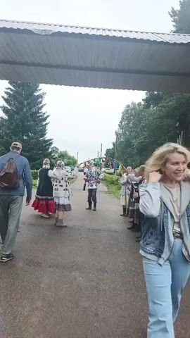 A group of people in traditional clothing are gathered on a road. A man in the foreground on the left is walking away from the camera. In the center of the road, a woman in a white and patterned outfit is holding a stick with ribbons. To her right, a man is playing an accordion, and several other people are standing in a line, also dressed in traditional attire. The environment appears to be outdoors, possibly in a rural setting, with trees and a cloudy sky visible in the background. There is an archway or overhead structure in the foreground, obscuring the top of the scene.