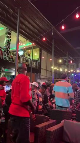 The video shows a crowded outdoor bar or restaurant at night. There are many people, some sitting at tables and others standing or walking around. A DJ and live musicians are performing on a small stage at the far end of the area. String lights are hung overhead, providing dim, colorful lighting. The atmosphere is lively and social. People appear to be enjoying the music, drinks, and company. The overall impression is a busy and festive night out.