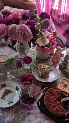 A table is laden with flowers, fruits, and desserts. In the background, a bouquet of pink and purple peonies dominates the scene, arranged in a clear glass vase. To the right of the peonies, a smaller arrangement of pink and white flowers sits in a white pot. Further right, a decorative golden object rests on a striped pink and white curtain. In the foreground, a woven placemat holds a plate with a slice of tiramisu, drizzled with chocolate sauce. To the left of the placemat, a bowl of raspberries and another bowl of blueberries are visible. At the bottom right, a whole crumble cake is presented, with a slice removed and a knife nearby. Two small vases, each holding a single purple flower, are placed to the left of the placemat. A glass of liquid and a white plate with what appears to be dried flowers are also on the table. The overall atmosphere is that of a delightful and abundant spread.