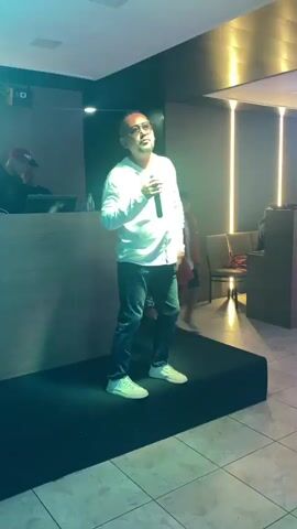 A man wearing a white collared shirt, blue jeans, and white sneakers stands on a raised platform and sings into a microphone. He has short hair and is wearing sunglasses. The platform is black and located in the center of a room with a tiled floor. Blue and white lights illuminate the scene, casting shadows. In the background, there is a DJ booth with two people behind it and several individuals standing or seated in the room, some of whom are partially visible and out of focus. The room appears to be a dimly lit indoor venue, possibly a club or restaurant, with decorative lighting on the walls. A young person wearing a red scarf draped over their shoulders walks past the platform in the background.