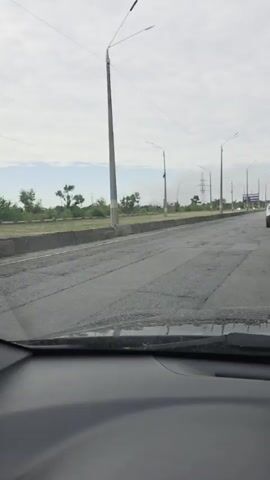 The video is filmed from inside a car, showing the view through the windshield. The road ahead is paved but shows significant wear and tear, with numerous potholes and cracks visible. To the left of the road, there is a concrete barrier and a grassy area with scattered trees. Several streetlights and power lines run along the right side of the road. In the distance, a billboard is visible, along with other cars and a power pylon. The sky is cloudy.