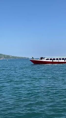 The video shows a red and white boat traveling on a body of water on a sunny day. The water appears to be a deep blue with gentle ripples. The boat has the word "FAJAR" visible on its side. On the front of the boat, one person is standing and another sitting. In the distance, there is land covered in greenery. The sky above is clear and blue.