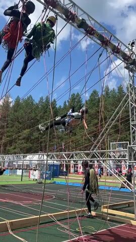 This video captures an outdoor obstacle course event, likely a competition, taking place on a sunny day with a clear blue sky and scattered clouds. The setting appears to be a sports complex or park, with a running track and trees in the background.

The primary subjects are several people navigating a challenging rope-based obstacle. Multiple individuals are suspended in the air, utilizing ropes and harnesses for support and movement. One person in a green outfit is positioned higher up on a metal structure, while another in a blue and white outfit is hanging upside down, appearing to be in the middle of a maneuver. Below them, another person in dark clothing is on the ground, seemingly preparing to ascend or having just completed a section. The complex arrangement of ropes, carabiners, and metal scaffolding forms the core of the obstacle.

The activity involves participants climbing, swinging, and maneuvering through a series of ropes suspended between metal frames. This is clearly a physically demanding challenge requiring strength, balance, and coordination. The presence of spectators and what looks like event tents in the background suggests a public event.

The time of day is likely midday or early afternoon, given the bright sunlight casting shadows. The weather is pleasant, suitable for an outdoor event. While no specific text is clearly legible in the video, the scene itself conveys a sense of athleticism and determination. The expressions on the faces of the participants, though not always clear, hint at focus and possibly exhilaration or exertion. The overall atmosphere appears energetic and competitive.