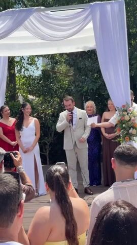 This video captures a wedding ceremony taking place outdoors under a white canopy. A man in a light-colored suit with a burgundy tie is speaking into a microphone, facing a woman in a white wedding dress with a high slit. The woman is smiling and looking at the man. To the left of the bride are two women in formal dresses, one in red and one in pink. To the right of the groom are two women in formal dresses, one in purple and one in brown, and a floral arrangement. In the foreground, several guests are visible, some holding up phones to record the event. The atmosphere appears celebratory, with guests clapping and cheering. The setting is outdoors with greenery and trees visible in the background, suggesting a sunny day.
