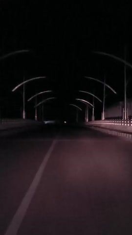 The video shows a dark, empty road at night, possibly a bridge, with streetlights lining both sides. The camera appears to be mounted on a moving vehicle. In the distance, there are faint lights, suggesting other vehicles or distant structures. The road has a white line marking the lane. The overall atmosphere is dark and deserted.