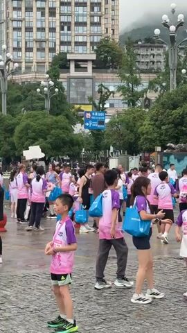 The video shows a gathering of people, many wearing matching pink and purple shirts, possibly after a race or event. Individuals of various ages are visible, including children and adults. Some people are carrying blue bags, and some are eating. The setting appears to be an outdoor public space, possibly a plaza or park, with buildings and trees in the background. The ground is wet, suggesting it may have rained recently. There are green tents set up in the background, possibly for event support or vendor stalls. The overall atmosphere suggests a post-event gathering.