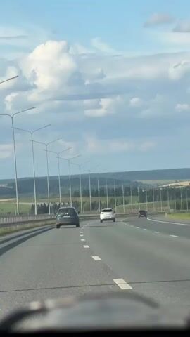 This is a vertical, eye-level, long shot of a highway on a sunny day with scattered clouds. The camera appears to be inside a vehicle looking out at the road ahead. Several cars are visible on the highway, moving in the same direction. The road has multiple lanes and is bordered by a row of tall streetlights on the left and a line of trees on the right. The landscape beyond the trees appears to be rolling hills and fields. The sky is blue with fluffy white clouds.