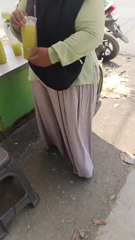 The video opens with a close-up of a person's hands holding a yellow object, possibly a drink or food item. The scene then shifts to a wider view, showing the person's lower body, wearing loose-fitting light brown pants and sandals. They are standing next to a green and white cart or stall. On the cart are two bottles of a light green liquid, one of which is partially empty, and a glass filled with the same liquid, with a red straw in it. Another person's hand, with a dark sleeve, is visible in the foreground, interacting with the cart or an object on it. The ground appears to be paved and has some debris, including leaves. The lighting suggests it is daytime, with shadows visible. The camera angle is low, looking up towards the people and the cart.