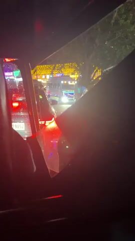 The video, shot at night, shows a busy street from inside a vehicle. The windshield is wet, possibly from rain. Numerous cars and auto-rickshaws are visible, their headlights and taillights creating a glowing scene. In the background, buildings are adorned with many small, yellow lights, resembling festive decorations. The overall impression is one of a vibrant, active urban environment at night.