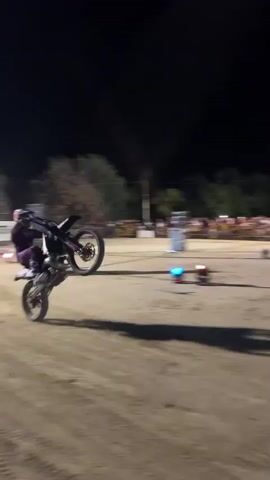 A nighttime motocross stunt show is underway. A person wearing purple gear rides a dirt bike, performing a wheelie. The rider is in motion, with the front wheel of the motorcycle in the air. Dust is kicked up from the ground, obscuring the view of the rear wheel in some shots. Spectators are visible in the background, separated by a fence. Lights illuminate the scene, and a large, dark structure, possibly a building or shipping container, is visible in the background of one shot. The moon is also visible in the night sky. The overall atmosphere suggests an outdoor event with a crowd gathered to watch the stunt performance.
