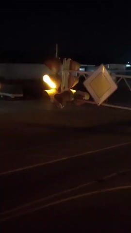 A traffic light and a diamond-shaped road sign are visible at night. The traffic light is positioned above the diamond sign, which appears to be a yield sign. The traffic light is mounted on a pole. The scene is dimly lit, with some ambient light from distant sources.