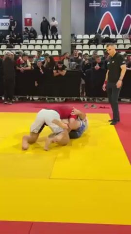 Two men are wrestling on a yellow and red mat in what appears to be a sports arena. The man on top is wearing a red shirt and white shorts. The man underneath is wearing patterned shorts. A referee stands nearby, watching the match. The area around the mat is filled with spectators sitting and standing, observing the competition. Banners and advertisements line the walls of the arena.