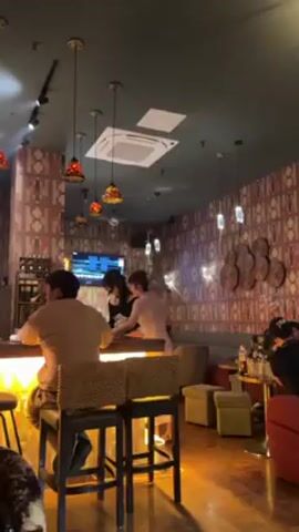 The interior of a bar is shown in this video. A lit bar with three patrons seated on stools is visible on the left side of the scene. One person stands behind the bar. A television hangs on the wall behind the bar. Seating areas with couches and tables are located in the right side of the scene. A second server walks from behind the bar towards the seating area. Patterned wallpaper covers the walls. The room is lit by a combination of overhead track lighting, pendant lights, and bar lighting.