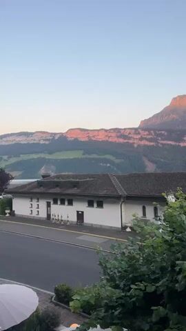 This is a vertical video capturing a serene waterside scene in Gersau, Switzerland, during the golden hour. The sun is setting, casting a warm, rosy glow on the majestic mountains in the distance. The sky is a soft gradient of blue and pink, with a sliver of the moon visible.

In the foreground, a cluster of lush green trees partially obscures a building with a distinctive yellow sign bearing the text "Ländi". The building appears to be a commercial establishment, possibly a shop or restaurant, with a traditional Swiss architectural style. Adjacent to this building, a paved road runs alongside the water.

Further into the scene, a calm lake stretches out, reflecting the soft light. A dark-colored car is parked near the water's edge, and a Swiss flag is prominently displayed on a flagpole. Another building, possibly a pavilion or cafe, with a dark, sloped roof, is visible on the right side of the frame. The overall atmosphere is peaceful and picturesque, suggesting a tranquil evening in a scenic Swiss town. The time of day is clearly evident from the lighting conditions. The weather appears to be clear and calm.