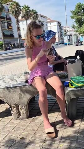 A woman with blonde hair in a ponytail, wearing sunglasses, a pink tank top, and pink shorts, sits on a concrete bench. She is holding and examining a small, brown object in her hands. To her right, a child wearing a blue and white patterned hat is seated on the same bench, looking at a phone. On the bench between them are several objects, including a green lunch bag, a white bag, and a bottle. The background shows a street with palm trees, buildings, and cars. The ground in front of the bench is paved with interlocking bricks.