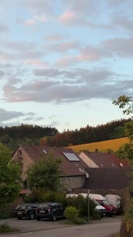 The video shows a row of buildings in a rural area under a partly cloudy sky. The buildings are mostly two-story structures with brown tiled roofs. Solar panels are visible on one roof. Several cars are parked along the street in front of the buildings. In the background, a hill covered with trees is partially illuminated by the late afternoon sun. A field of golden crops is visible at the top of the hill. The foreground is partially obscured by trees and bushes.