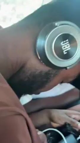 Here's a description of the video:

The video shows a man in the backseat of a car wearing a brown shirt, a black cap, and silver JBL headphones. He is looking down at a phone or device in his hands, and a charging cable is visible. Other people are in the car with him. The car is moving.