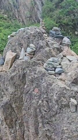 This is a vertical video of a rocky embankment next to a body of water. There are several cairns, which are stacks of stones, placed on the rocks. The rocks are various shades of gray and brown, and some have moss growing on them. The water appears clear and shallow, with a rocky bottom visible. The background consists of a steep, vegetated cliff face. The video is shot from a close-up perspective, focusing on the details of the rocks and cairns. There are no people or animals visible in the video. The lighting suggests it is daytime, possibly overcast.