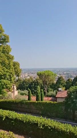 The video opens with a panoramic view of a town or city in the distance, seen from a hilltop vantage point. A clear blue sky dominates the upper portion of the frame. In the foreground, lush green trees and hedges line a stone wall, creating a sense of height and perspective. As the camera pans to the right, a quaint building with a red-tiled roof comes into view, partially obscured by dense foliage. Further along the wall, the ruins of another structure, also covered in ivy, are visible. The camera continues its sweep, revealing a more prominent, light-colored building with a pitched roof and multiple windows. This building sits atop a gentle slope, with a pathway and more greenery leading up to it. The scene then shifts to a road curving through a landscape dotted with tall cypress trees and other dense vegetation. The road itself is bordered by a stone wall on one side and a lush green hedge on the other. Finally, the video concludes with a view of a medieval-style stone tower, part of a larger castle structure, rising against the blue sky. A curved stone wall and pathway lead towards it, and two people, an adult and a child, are walking along the path, partially obscuring the lower part of the tower. The overall impression is one of a scenic and historical location, likely in a Mediterranean region, bathed in bright sunlight.