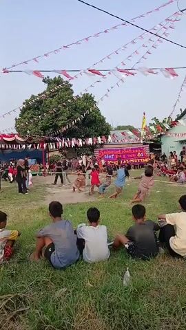 A group of children are playing a tug-of-war game outdoors. They are on a grassy field, and there are spectators sitting on the grass watching them. In the background, there is a tent with decorations, including Indonesian flags. The sky is clear and the sun is out. The children are all wearing casual clothing and appear to be enjoying themselves.