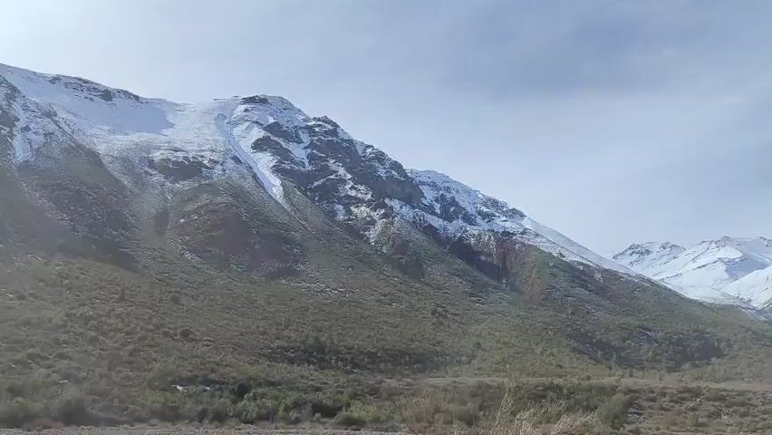 The video shows a wide aerial shot of a mountainous landscape. The foreground is filled with low-lying vegetation and bare trees. The middle ground consists of a large, steep mountain slope covered in patches of snow and dry earth. In the background, several other snow-capped mountain peaks are visible against a bright, overcast sky. The camera pans slowly to the right, revealing more of the snowy mountain range. There are no people or man-made objects visible in the video. The environment appears to be cold and possibly at a high altitude.