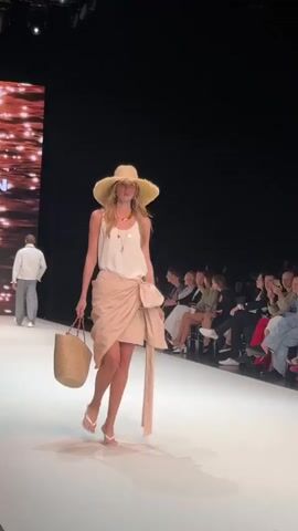 A fashion show is taking place with a model walking down a runway. She is wearing a large straw hat, a white tank top, and a tan wrap skirt. She is carrying a straw purse in her left hand and wearing white heeled sandals. In the background, an audience is seated on the right side of the runway, and a large screen displaying abstract images is visible on the left.