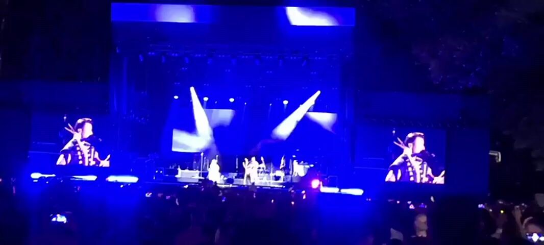 A wide shot of a concert stage with bright blue lighting. Two large screens on either side of the stage display a close-up of a male singer in a decorative jacket, holding drumsticks to his mouth. The main stage features several musicians, including a drummer and a person playing a cello, all bathed in blue light from above and from large, bright white spotlights. A large crowd of people is visible in the foreground, some holding up their phones to record. The overall atmosphere suggests an outdoor evening concert.