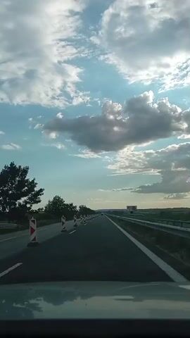The video shows a view from inside a vehicle traveling on a highway. The road is asphalt and has clear white lane markings. Orange and white construction cones are lined up on the left side of the road, indicating a work zone. The sky is filled with dramatic clouds, with patches of blue sky visible and the sun peeking through. In the distance, there are some trees and a billboard on the right side of the highway. The overall impression is of a journey through a partly cloudy landscape.