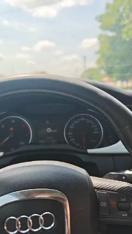 The video captures an interior view of an Audi car being driven in Preslaven, Bulgaria, during daytime under sunny skies with scattered clouds. The primary focus is on the black leather steering wheel, emblazoned with the Audi rings logo, and the instrument cluster behind it.

At the beginning of the video, the car is stationary, displaying "0 km/h" on the digital readout. The odometer shows 473,118 km, and a trip meter reads 178.7. An orange warning light (possibly engine check) and a low fuel indicator are visible on the dashboard. As the driver's right hand grips and turns the steering wheel, the car begins to move. The speed increases to 31 km/h and then to 38 km/h, indicated on both the analog speedometer and the digital display. A tachometer shows engine RPMs.

Through the windshield, a bright sky, trees, and glimpses of buildings and power lines suggest a residential or rural road. A partial view of the car's infotainment screen shows "FM radio," "CITY," and "Pop" selected. The driver's right hand is prominently visible, manipulating the steering wheel, and their foot/leg in light-colored footwear can be seen in the bottom right corner of the frame. Music is audible in the background throughout the video.