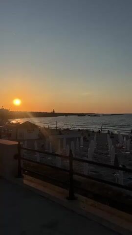 A vertical video captures a sunset over a beach. The sun, a bright orange orb, is positioned on the left side of the frame, casting a warm glow across the sky and water. The sky transitions from a pale blue at the top to a soft orange near the horizon. The sea is calm with gentle waves rolling onto the shore. A beach is visible with numerous rows of white beach umbrellas and lounge chairs. A few people are scattered across the beach and in the water. In the foreground, a dark railing separates the viewer from the beach scene. The video is captured from a slightly elevated perspective.