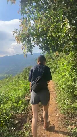 The video opens with a person, seen from behind, walking on a narrow dirt path. They are wearing a dark short-sleeved top, dark shorts, and light-colored sandals, with a black fanny pack strapped around their waist. Lush green foliage lines the path to their right, and a vast valley with distant mountains under a partly cloudy sky stretches out to their left. The camera then pans left, revealing a breathtaking panoramic view of the mountainous landscape. Rolling hills are covered in extensive, intricate terraced fields, some appearing green with crops, while others are a reddish-brown, possibly recently tilled or flooded. The scene is bathed in natural daylight, suggesting late afternoon or early evening. The overall setting depicts a serene and picturesque rural environment, characteristic of mountainous agricultural regions in places like Lao Chai, Vietnam. No visible text is present in the video.