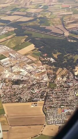 The video shows an aerial view from an airplane window of a town and its surrounding agricultural landscape. The town is densely populated with houses and buildings, and there are some industrial areas and roads visible. The landscape is a patchwork of fields, with some wooded areas and wind turbines in the distance. The lighting suggests it is daytime, likely with clear skies. The camera angle is angled downwards and slightly to the side, providing a perspective from the aircraft. There are no people or discernible actions visible within the frame. The video appears to be a continuous shot with no significant changes in the scene.