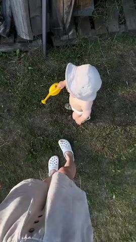 The video shows two people outdoors, likely in a rural setting in Krivandino, Russia. The first segment focuses on a baby on its hands and knees on grass, with a yellow watering can nearby. An adult's legs, wearing white Crocs and light-colored shorts, are visible in the foreground. The second segment is a selfie-style shot from a different adult, who is wearing a black bikini top and white shorts. This person is smiling and looking at the camera. Behind them, a wooden building with windows and stacked wood can be seen. The sun appears to be low in the sky, suggesting it is either early morning or late evening. The weather seems pleasant and sunny. The overall mood is relaxed and casual, with a focus on outdoor activity. There is no visible text in the video.