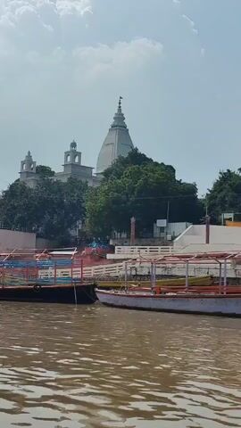 A wide shot captures a serene river scene with a large, white, multi-domed temple as the focal point in the background. The temple is flanked by lush green trees and a stone wall. In the foreground, several boats are moored on the muddy brown water. Some of the boats appear to be passenger vessels with colorful railings and seating. The sky is partly cloudy, with a few white clouds visible against a pale blue backdrop. The overall atmosphere is peaceful and tranquil.