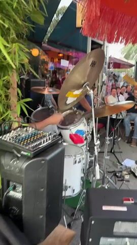 A medium shot shows a drummer playing a white drum kit. The drummer is wearing a black t-shirt and glasses. They are using drumsticks to hit the snare drum and cymbals. To the left of the drum kit is a black audio mixer. In the background, there are people and a red fringed umbrella. The scene appears to be outdoors, possibly at a cafe or restaurant.