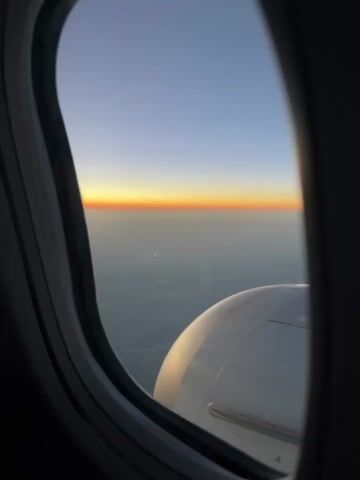 The video shows a view from an airplane window during sunset. The horizon displays a gradient of orange and yellow against a clear blue sky. Below the horizon, the landscape is obscured by a layer of clouds or haze, with faint indications of the earth's surface visible. A portion of the airplane's wing and engine cowling is visible in the lower right corner of the frame. The window frame is dark and slightly blurred.