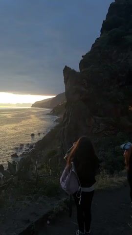 A young person stands on a cliffside overlooking a body of water, likely the ocean, during what appears to be dusk or a time with low light. They are wearing a dark top and leggings, with a light-colored backpack. The ocean reflects a dim, golden light from the setting sun on the horizon. To the person's right is a rocky cliff face covered in vegetation, and in the foreground are cacti. The sky is overcast with dark clouds, and the scene is generally dark and moody.