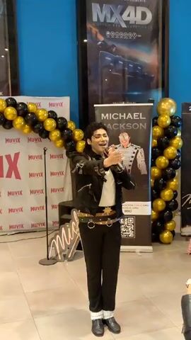 A man dressed as Michael Jackson is performing on a stage. He is wearing a black jacket with silver detailing, black pants, and white socks with black shoes. He is holding a microphone and appears to be singing. There are balloons behind him, some black and some gold. There is a banner with Michael Jackson's name on it. The floor is tiled and the walls are blue.