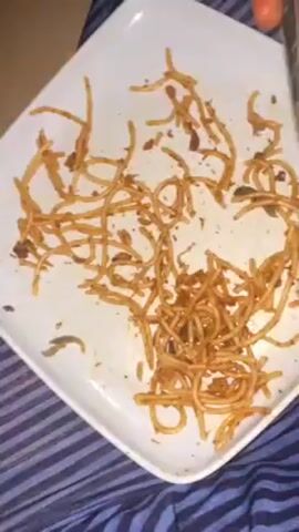 A close-up shot shows a white square plate with a scattering of spaghetti.  A fork is used to gather and move the pasta. The spaghetti is coated in a reddish-brown sauce and some dark green specks are visible. The plate rests on a blue and white striped cloth.  The pasta is being arranged on the plate, suggesting an intention to consume it.