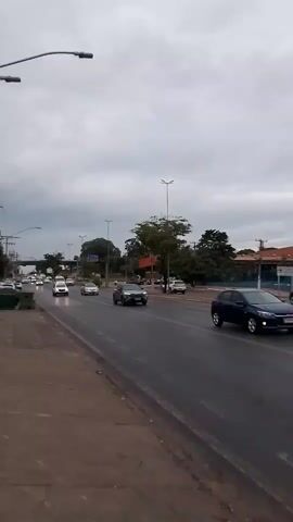 A video shows a street scene with cars and trucks driving on a wet road under a cloudy sky. A person is walking on the sidewalk on the left side of the road. A green trash can is visible on the sidewalk. There are trees and buildings in the background. A large red truck with "ÁGUIA SUL" written on its side is driving in the right lane. Several other vehicles are visible on the road, including cars, a white pickup truck, and a white van. The overall lighting suggests it is daytime, possibly late afternoon or early morning.